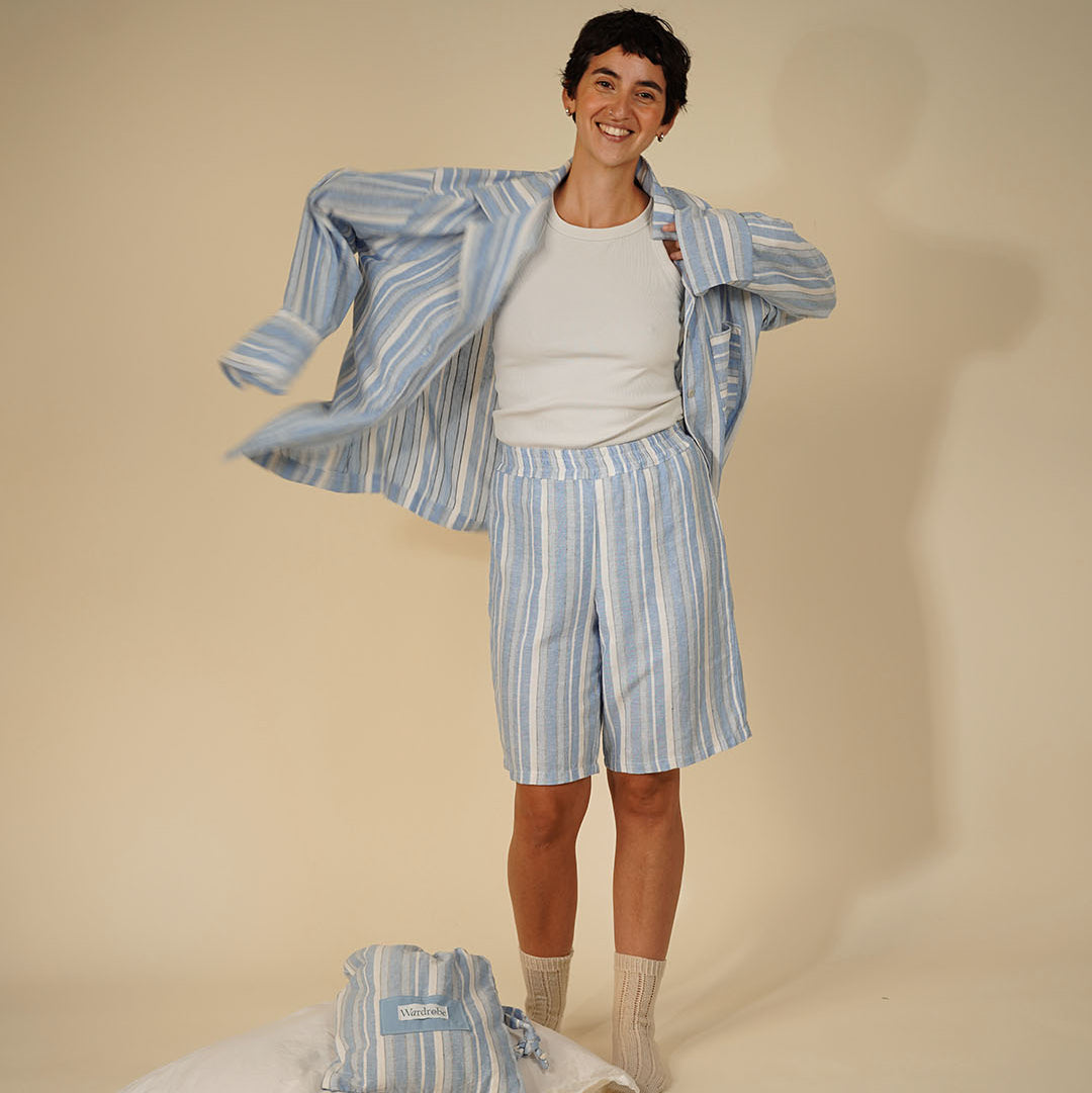 A smiling man models a light blue and white striped pajama set, including a long-sleeve shirt and shorts, over a white t-shirt. A matching folded pajama set rests on a white pillow on the floor next to him, set against a solid beige studio background.