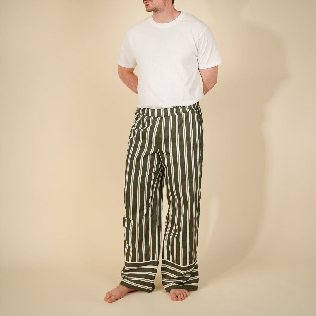 A man stands barefoot against a plain beige studio background, wearing a plain white short-sleeve t-shirt and loose vertical green-and-white striped pants.