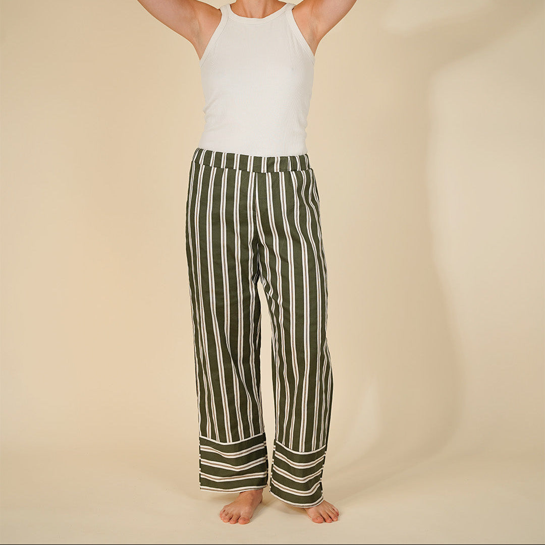 A model stands barefoot against a warm beige studio background, wearing a white sleeveless top and wide-leg trousers with vertical olive green and off-white stripes. The model has short dark hair and is holding it up with both hands.