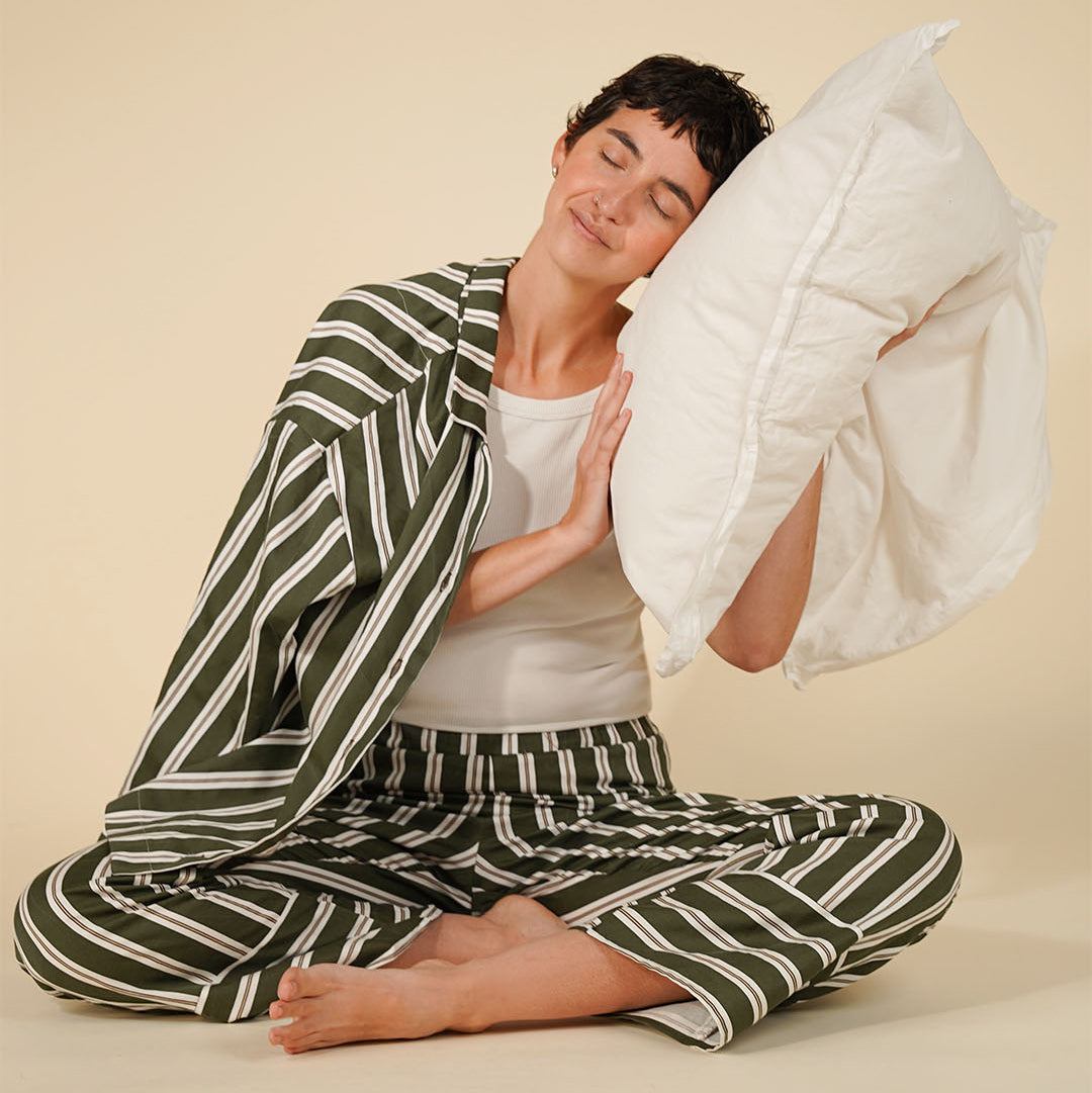 A woman sits cross-legged on a beige studio floor wearing a matching green and white striped pajama set, with a white pillow against her head.