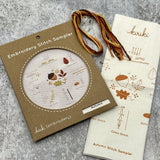 Kraft board packaging with cream cotton stitch sampler featuring an acorn and leaves and hank of cotton embroidery floss in autumn color palette