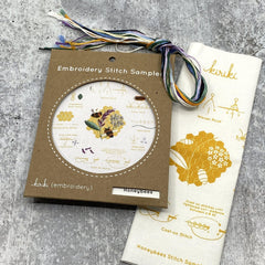 Kraft board packaging with cream cotton stitch sampler with honey bees and honeycomb and hank of cotton embroidery floss in summer color palette