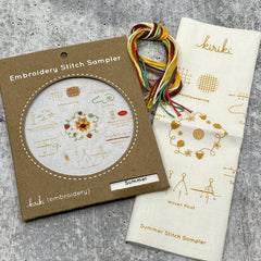 Kraft board packaging with cream cotton stitch sampler featuring a sunflower and strawberries  and hank of cotton embroidery floss in summer color palette